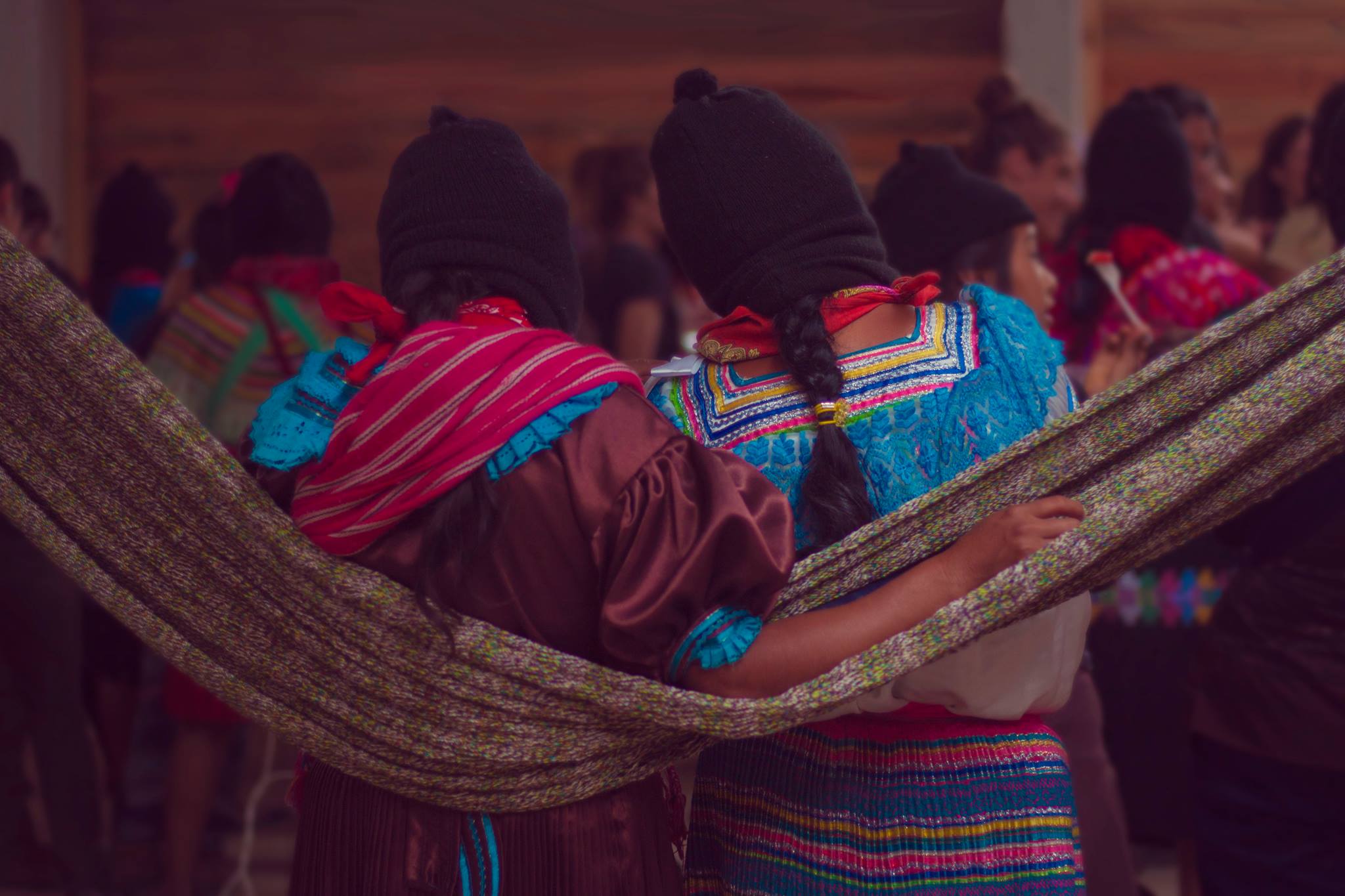 Dos mujeres se abrazan por la espalda mientras escuchan atentas hacia adelante. Visten sus trajes tradicionales