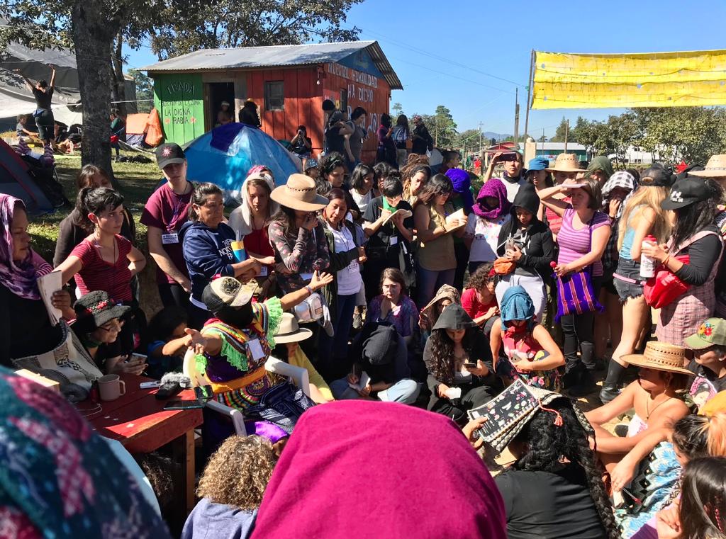 Mujeres se colocan en círculo para escuchar atentamente a una mujer sentada. Viste traje tradicional y pasamontañas y tiene los brazos abiertos. Mira: feminismos y democracias